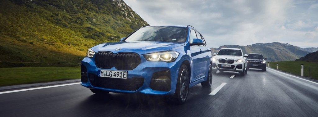 Un BMW X1 bleu circulant sur une route avec d'autres SUV, entouré de collines verdoyantes et sous un ciel nuageux.